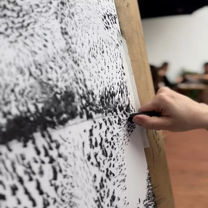 Mão desenhando com carvão em papel preso ao cavalete durante aula prática de artes visuais