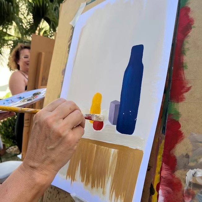 Mão segurando pincel e pintando garrafa azul e formas coloridas em tela durante oficina de artes